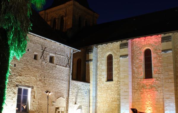 Une photo de nuit à l'extérieur du Prieuré Saint-André éclairé. Le public écoute les musiciens à côté d'un palmier.