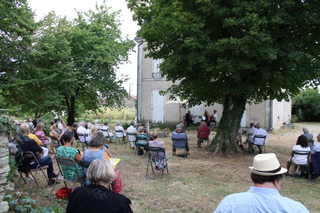 Une photo d'un concert à la Maison Tassin. Du public assis en plein air écoute des musiciens.