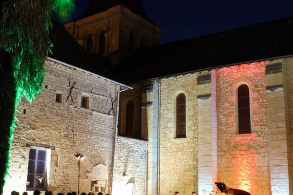 Une photo de nuit à l'extérieur du Prieuré Saint-André éclairé. Le public écoute les musiciens à côté d'un palmier.