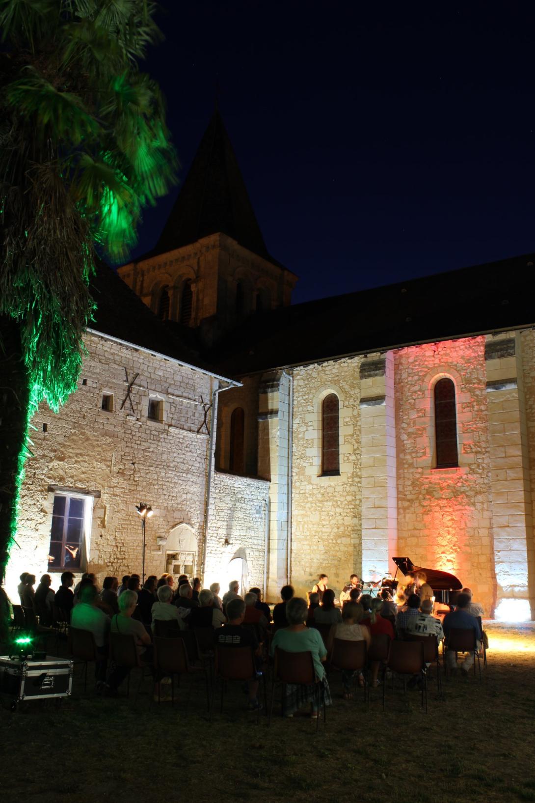 Une photo de nuit à l'extérieur du Prieuré Saint-André éclairé. Le public écoute les musiciens à côté d'un palmier.